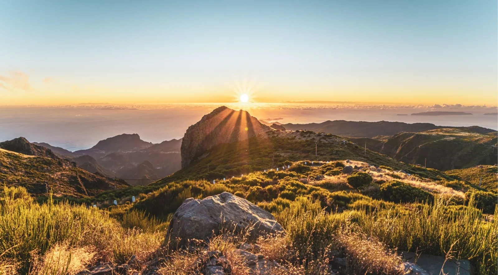 The sun breaking over a mountain ridge above the Atlantic, seen from a grassy plateau near Achada do Teixeira