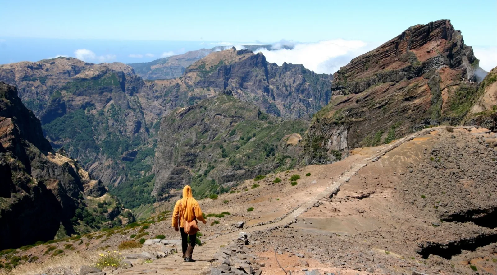 A hiker in an orange jacket walking down the stone path from Pico do Arieiro toward the ridge