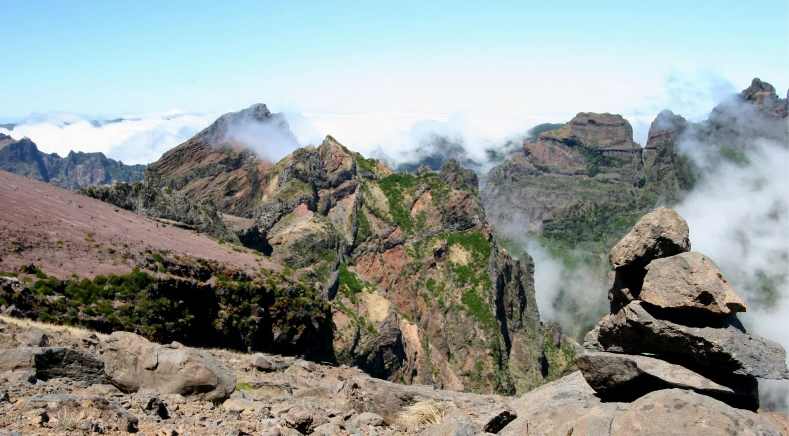 Red volcanic peaks rising above a solid white cloud layer, with a small stone cairn in the foreground