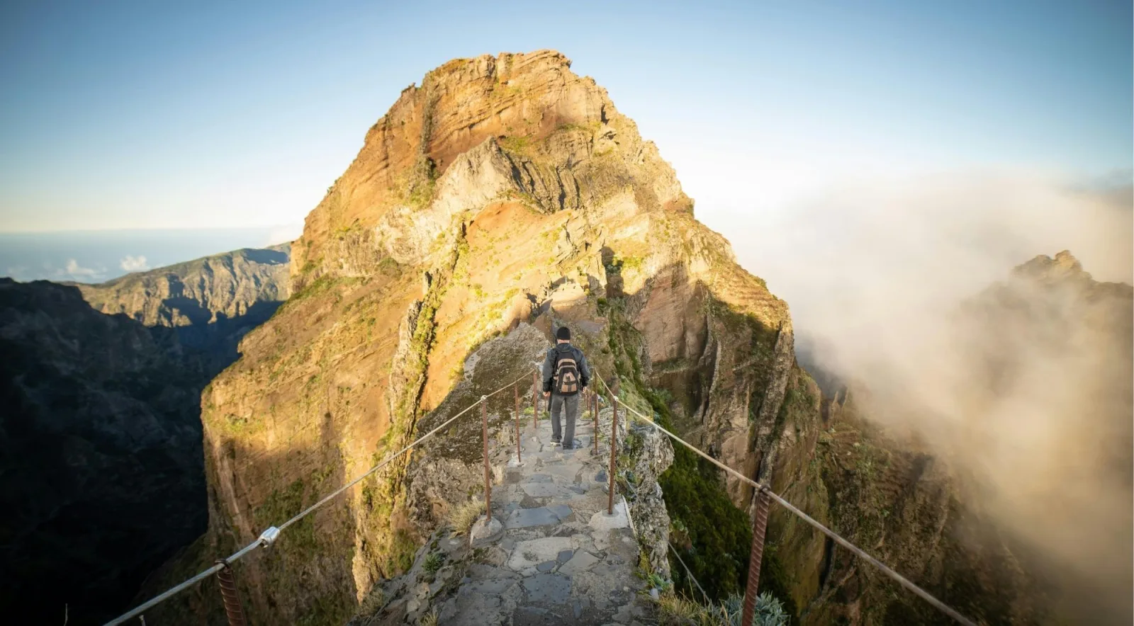 A hiker with a backpack crossing the cable-railed stone stairway on the PR1 ridge, mist rising on the right