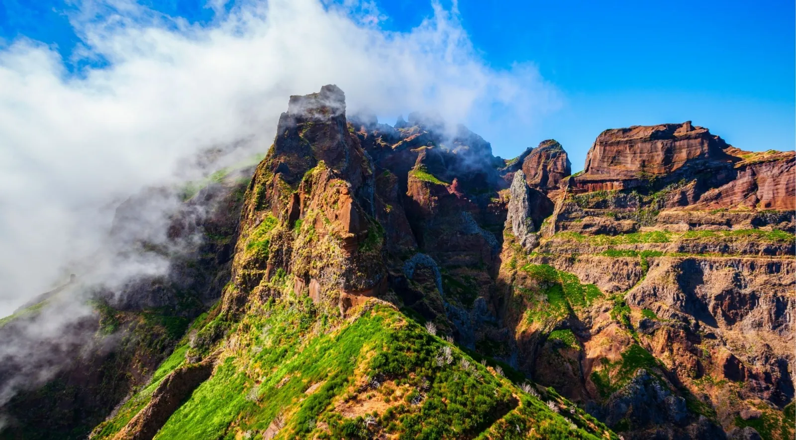 A green and red rock ridge under a bright blue sky with cloud spilling over the crest