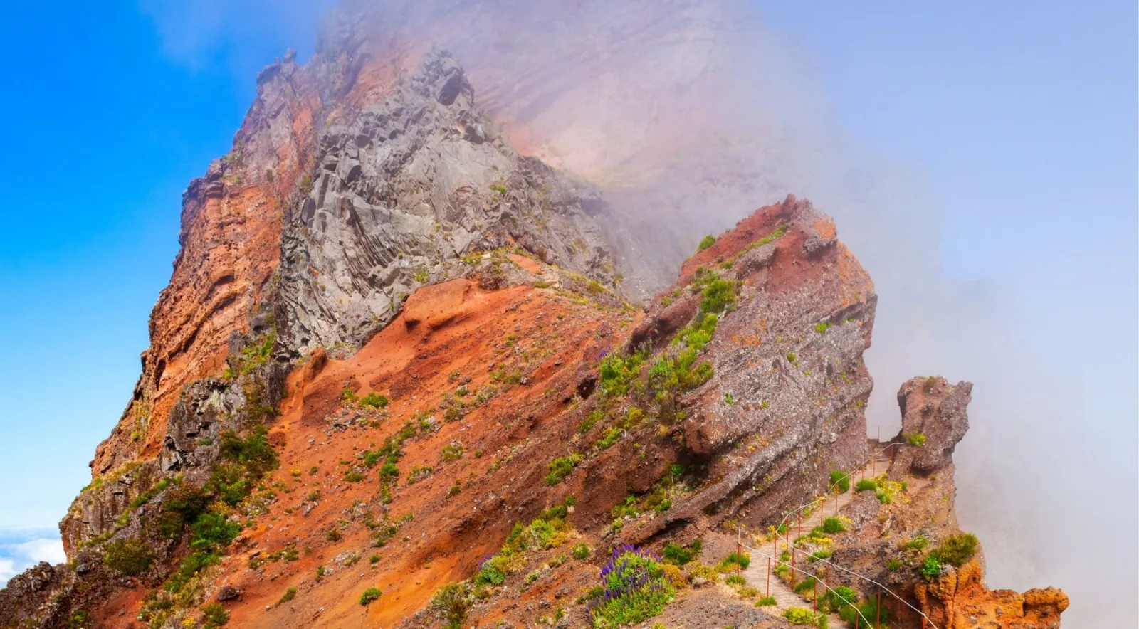 Orange volcanic rock along the PR1 with the railed path traversing the slope into thick mist